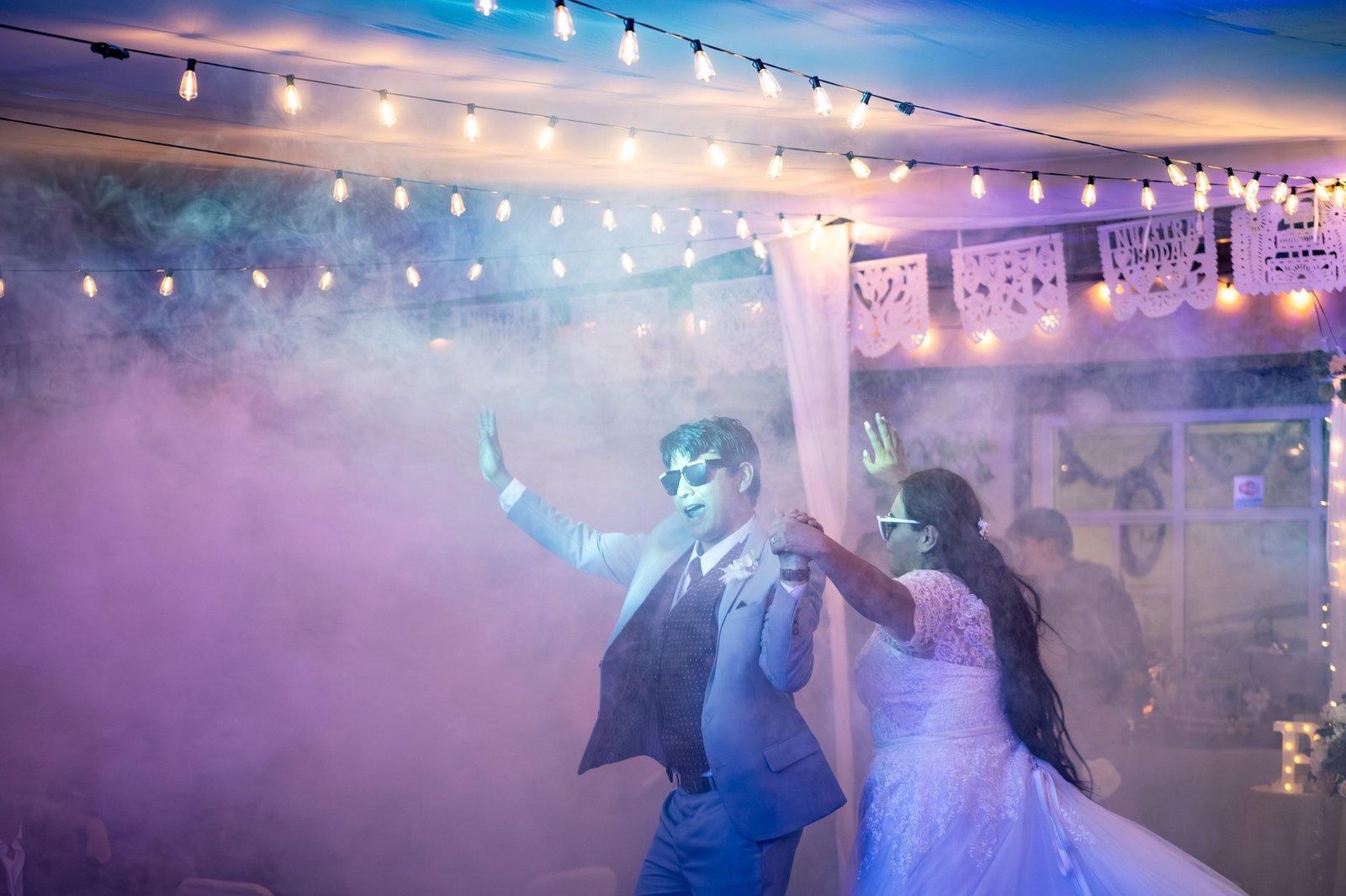 Pareja bailando entre humo de colores bajo series de luces y papel picado